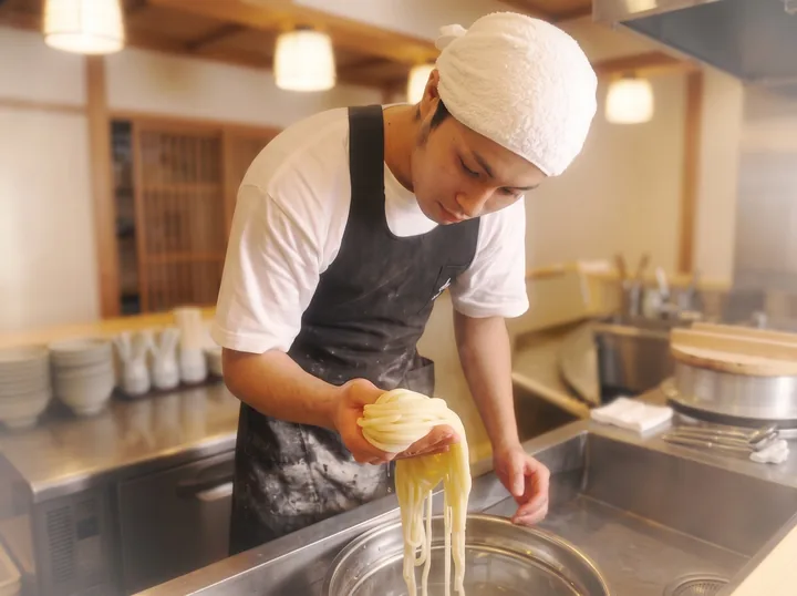 若き日の谷 和幸が締めたての麺を手に取る仕事姿（イメージ）